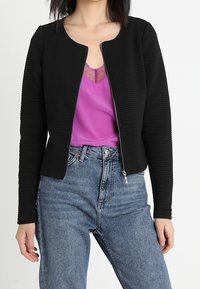 Svart strukturerad jacka med rund halsringning, frontdragkedja, lagd över en lila topp, tillsammans med ljusblå högmidjade jeans.