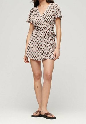 Frau in einem kurzen, gemusterten Wickelkleid mit kurzen Ärmeln und braunen Sandalen, die vor einem neutralen Hintergrund steht.