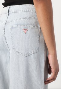 Stripete lyseblå denimjeans med en trekantet rød og hvit logo-patch på baklommen, med standard lommer og syddetaljer.