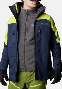 Giacca da sci blu e verde lime con zip frontale, multiple tasche e rivestimento in pile grigio testurizzato sotto. Guanti neri.