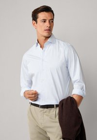 Muškarac nosi bijelu košulju sa tankim plavim kockastim uzorkom, bež hlače, crni pojas, drži presavijeni tamnosmeđi džemper u jednoj ruci.