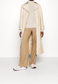 Long beige trench coat with gold buttons over a light beige knit sweater, paired with tan wide-leg trousers and white sneakers with red accents.