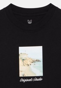 Μαύρο βαμβακερό μπλουζάκι με φωτογραφία ακτής σε μπεζ και ανοιχτό μπλε τόνους, με την κεντημένη ένδειξη "Originals Studio" σε λευκή γραμματοσειρά.