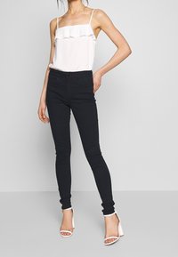 Černé skinny džíny s vysokým pasem, vyrobené z denimu, spárované s bílým tílkem s volánky. Obuv zahrnuje bílé sand ály na bloku.