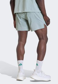 Άνδρας που φοράει ανοιχτοπράσινα αθλητικά σορτς, λευκές κάλτσες Adidas και λευκά παπούτσια τρεξίματος, στέκεται με τα πόδια ορατά από πίσω σε απλό φόντο.