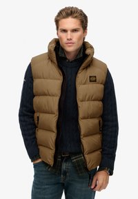 Gilet imbottito marrone con alto colletto, texture trapuntata orizzontale e tasche con zip, indossato sopra un maglione blu a trecce e una camicia a quadri.