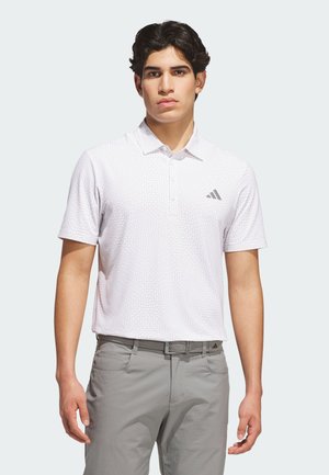 Junger Mann, der ein weißes kurzärmeliges Polo mit feinem Muster und graue Hose trägt, steht mit neutralem Gesichtsausdruck vor hellem Hintergrund.