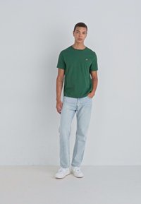 Tricou verde din bumbac, cu mâneci scurte, guler ușor rotund și un logo mic. Asortat cu blugi ușor albaștri și teniși albi.
