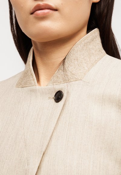 Κοντινό πλάνο ενός ατόμου που φοράει μπεζ παλτό με υφή, με ανεβασμένο γιακά και ένα μαύρο κουμπί κοντά στον λαιμό.