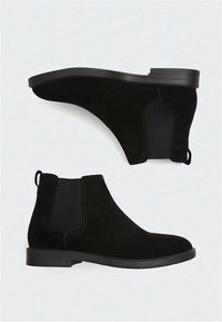 Une paire de bottes Chelsea en suedine noire, avec des panneaux élastiques sur les côtés et des languettes, présentée sur un fond clair, une botte à la verticale et l'autre sur le côté.