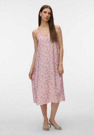 Mujer con un vestido midi sin mangas rosa con flores, tirantes finos y sandalias de tacón gris claro, de pie frente a un fondo liso.