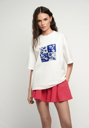 Kobieta z długimi włosami ubrana w biały oversize t-shirt w niebieskie kwadraty z motywem kwiatowym oraz czerwoną plisowaną mini spódniczkę, stojąca na jednolitym tle.