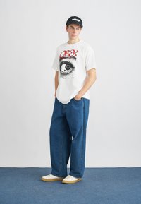 T-shirt branca com um gráfico de olho preto e texto vermelho, combinada com jeans azuis largos e tênis brancos com sola bege.