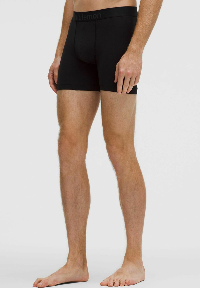 Fekete, testhez simuló boxer alsónadrágot viselő férfi mezítláb áll egy sima fehér háttér előtt.