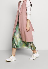 Dlouhá růžová vesta bez rukávů přes bílý žebrovaný svetr, doplněná splývavou zelenou květovanou sukní a bílými teniskami.