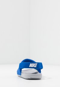 Sandales Nike bleues avec une semelle texturée grise et une sangle en tissu. "Nike" imprimé en blanc sur la sangle. Design simple et à dos ouvert.