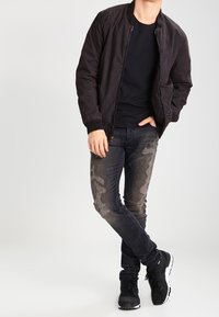 Chaqueta bomber negra sobre una camiseta negra, combinada con jeans oscuros desgastados con parches claros y zapatillas deportivas negras.