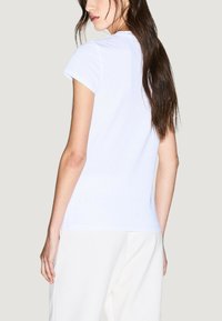 Camiseta blanca ajustada de manga corta y cuello redondo, hecha de algodón. La vista trasera muestra una textura suave y detalles de costura sutiles.