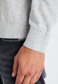 Manche de pull gris clair en maille montrant la texture côtière du poignet ; portée avec un jean en denim gris foncé, une main reposant contre le tissu.