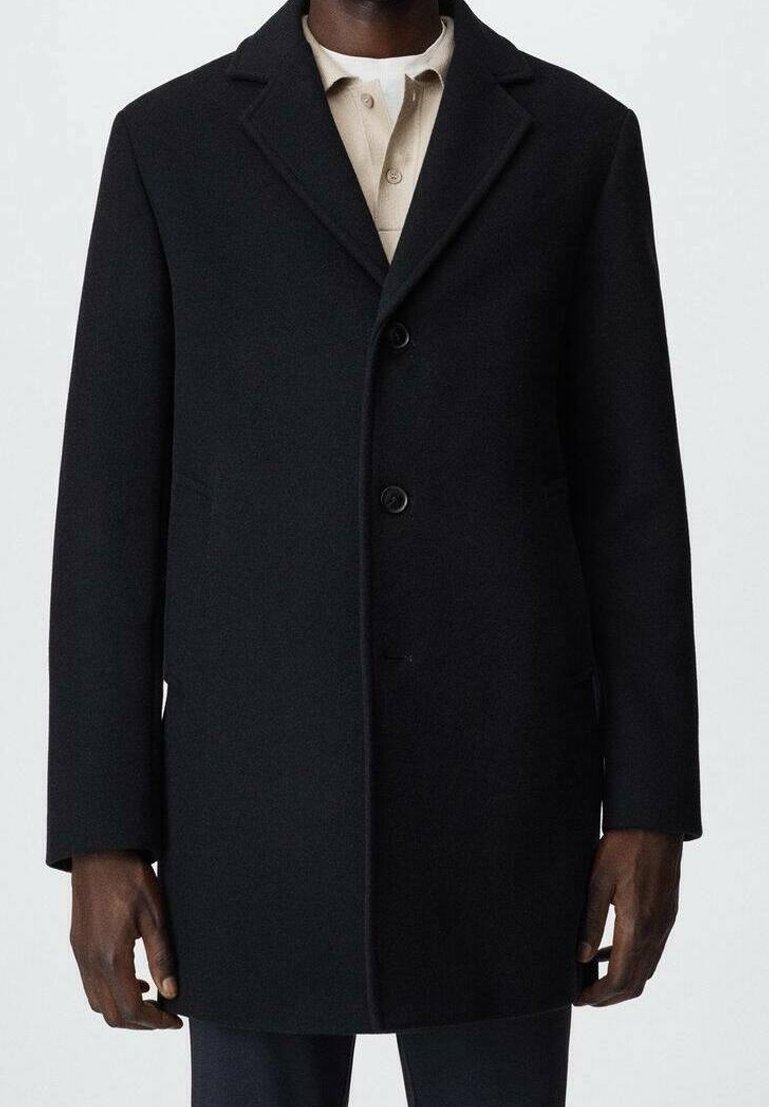 Manteau noir pour homme longueur genou avec col cranté et deux boutons, porté sur une chemise beige à col.