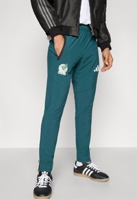 Türkiz atlétikai nadrág elasztikus derékpánttal, oldalzsebekkel és Mexikó hímzett logójával. Fekete Adidas csíkokkal és cipzáras mandzsettákkal rendelkezik.