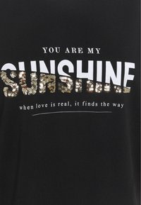 Tissu noir avec texte en sequins blancs et dorés disant « YOU ARE MY SUNSHINE » et, en dessous, « when love is real, it finds the way ».