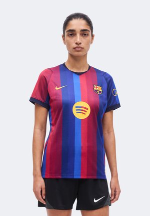 Femme aux cheveux foncés tirés en arrière, portant un maillot de football rayé bleu et rouge du FC Barcelone et un short noir Nike, debout, face à l'avant.