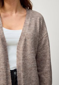 Długi kardigan z otwartym przodem w jasnobrązowym dzianinowym materiale o miękkiej fakturze, z ribbowanymi krawędziami, nałożony na biały top.