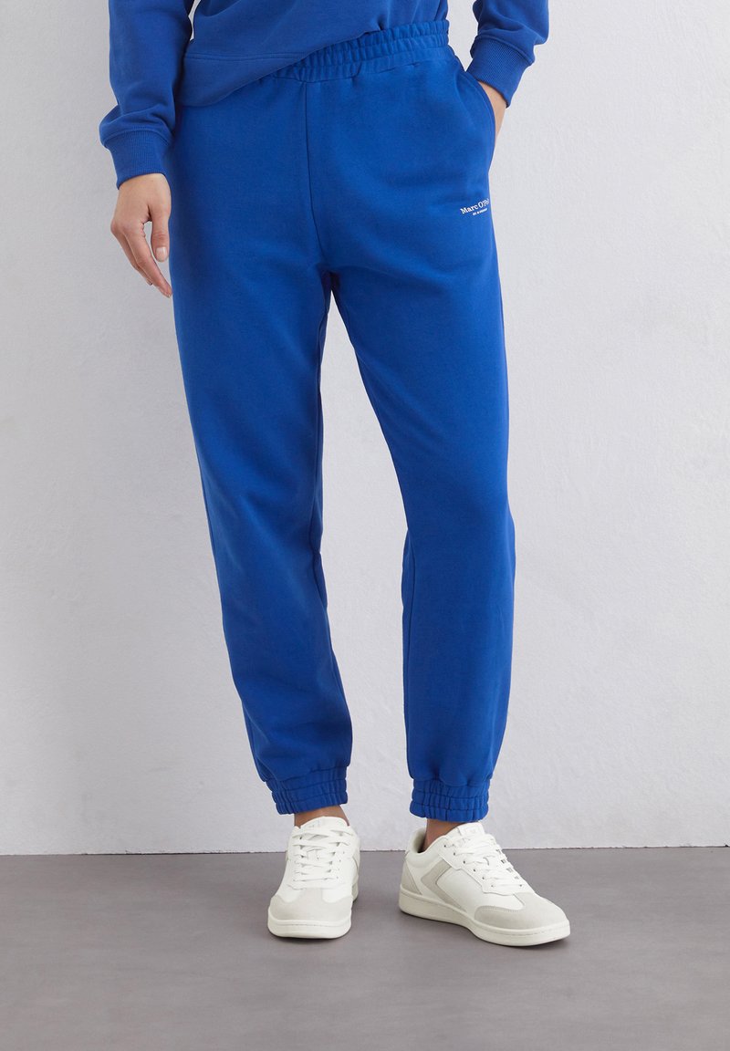 Blauwe katoenen sweatpants met een elastische tailleband, afgeboorde uiteinden en witte logodetails. Gecombineerd met witte sneakers op een grijze ondergrond.