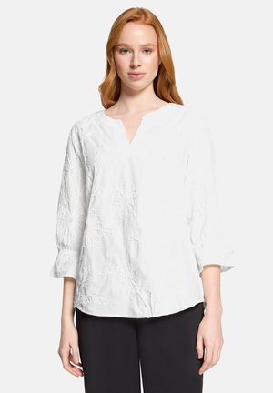 Ung kvinde med langt rødt hår iført en hvid bluse med blomstret broderi og 3/4-ærmer samt sorte bukser foran en ensfarvet baggrund.