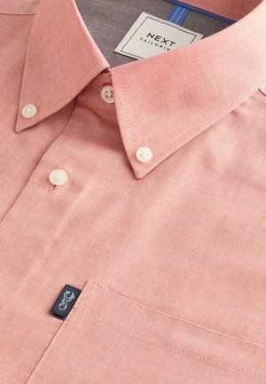 Light pink button-up shirt made of textured fabric, featuring a pointed collar, white buttons, and a small black logo tag on the pocket.