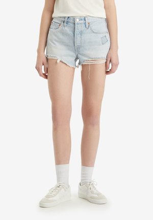 Jeune personne portant un short en denim bleu clair usé, des chaussettes blanches, des baskets blanches et un haut blanc rentré, debout sur un fond uni.