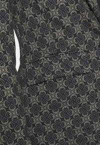 Černá látka s květinovým geometrickým vzorem v zelené a bílé barvě. Obsahuje detail sklopené kapsy na levé straně, hladká textura.