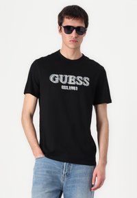 Juodas medvilnės marškinėlis su žodžiu "GUESS" ir "EST. 1981" spalvingu šriftu. Dėvima su šviesiai mėlynomis džinsų kelnėmis ir juodais saulės akinių.