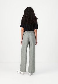 Personne aux longs cheveux bouclés portant un haut noir à manches courtes, un pantalon gris à jambes larges et des baskets blanches, debout devant un fond blanc uni.