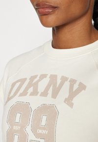 Gaiša krēmkrāsas džemperis ar "DKNY" logotipu izbalējušā bežā un "89" tekstūrā, ar ribotu apkakli un gludu audumu.