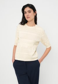 Πλεκτό κρεμ πουλόβερ με διάτρητο μοτίβο σε σχήμα ρόμβου, ριμπ λεπτομέρειες και κοντά μανίκια. Το σχέδιο διαθέτει λαιμόκοψη με κυματιστά άκρα και υφασμένες λωρίδες.