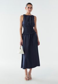 Abito senza maniche di colore blu navy con vita arricciata e orlo a volant, realizzato in tessuto leggero. Abbinato a una piccola borsa a triangolo bianca e sandali con cinturini.