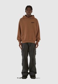 Felpa cappuccio marrone con tasca frontale e logo; pantaloni cargo neri con più tasche; indossata con sneakers scure.