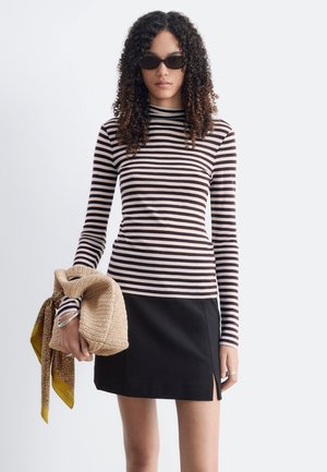 Femme aux cheveux bouclés portant des lunettes de soleil noires, un haut à manches longues rayé noir et blanc, une jupe noire, tenant un sac beige avec une écharpe à motifs.