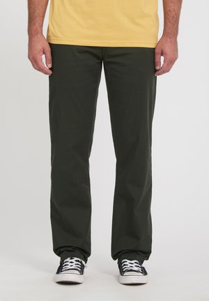 Homme portant un pantalon droit vert foncé, des baskets noires et blanches, et une chemise jaune sur un fond uni.