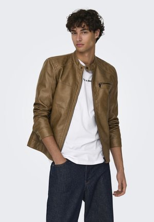 Giacca di pelle marrone con tasca sul petto con zip, colletto rialzato e dettagli a coste sulle maniche, indossata sopra una t-shirt bianca e jeans blu.