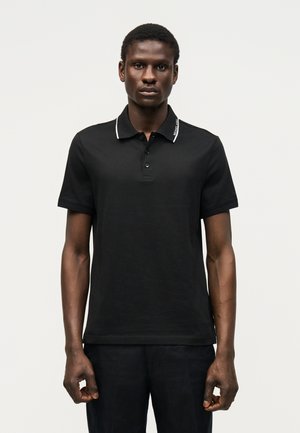 Fekete férfi, aki fekete, testre simuló galléros pólót visel, fehér szegéllyel a gallérnál, egyszerű világos háttér előtt áll.