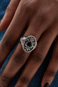 Zilveren ring met een hartvormige zwarte steen, sierlijke bloemengravures en getextureerde zijkanten, gedragen aan een hand met een natuurlijke huidskleur.