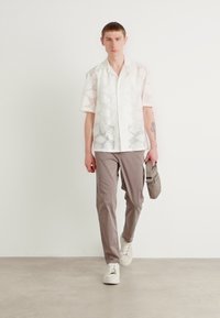 Witte kortärmedru blouse met een subtiel patroon, gecombineerd met lichtgrijze broeken en witte sneakers. Draagt een kleine grijze tas.