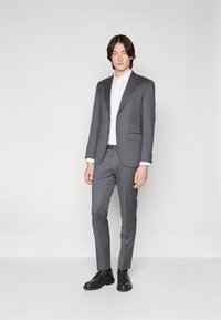 Grijze getailleerde pak met een smalle pasvorm, gecombineerd met een witte overhemd met knopen en zwarte leren schoenen. Soepele stof met strakke lijnen en minimale details.