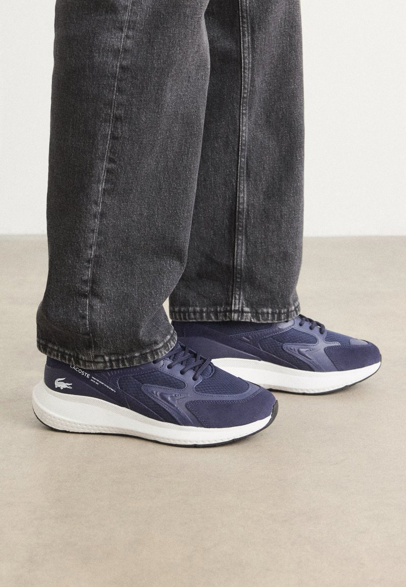 Marineblauwe sportschoenen van mesh en suede, met een witte zool en het Lacoste-logo op de zijkant, gedragen met donkergrijze jeans.