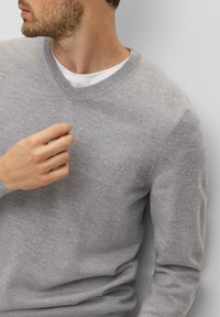 Pull en tricot gris avec un design en V. Présente un logo BOSS subtil sur la poitrine. Le matériau semble lisse et tricoté fin. Chemise blanche en dessous visible.
