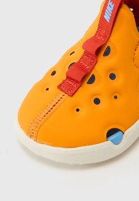 Oransje gummisko med ventilasjonshull, rød stropp og hvit såle. Har en blå aksent nær hælen og broderte detaljer.