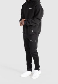 Sweat à capuche et pantalon de survêtement noirs en tissu doux et texturé. Le sweat à capuche présente une poche avant et un logo brodé ; le pantalon arbore un logo similaire.